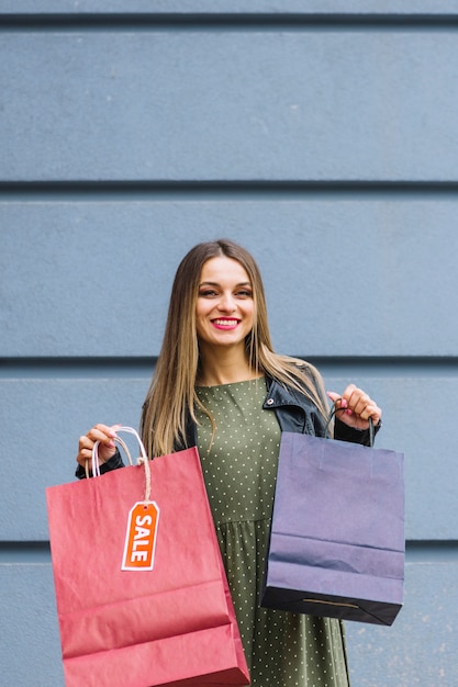 紫と赤の買い物袋を保持している壁に立っている笑顔の若い女性