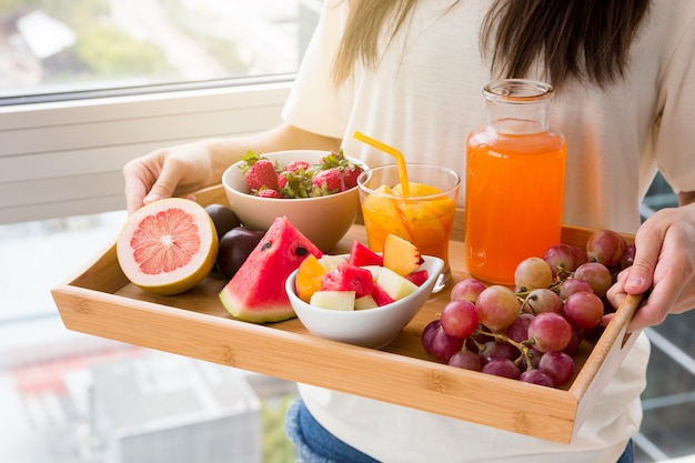 Smiling young woman standing against window holding tray of fruits and juice