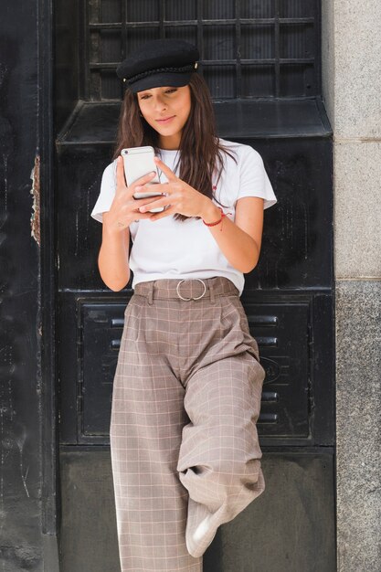 Smiling young woman standing in front of black painted wall texting message on smartphone