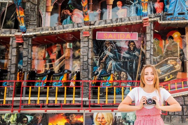 Smiling young woman standing in front of haunted house fun ride holding camera