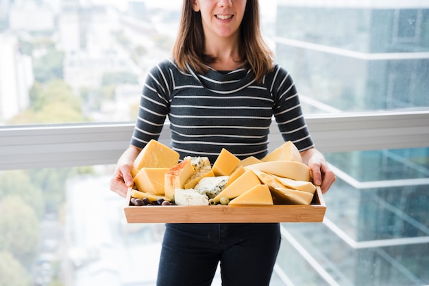 Free photo smiling young woman standing in front of window holding cheese in wooden tray