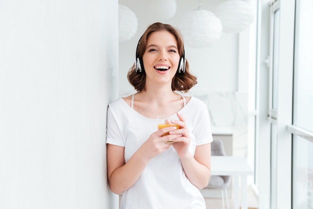Smiling young woman standing near window drinking juice