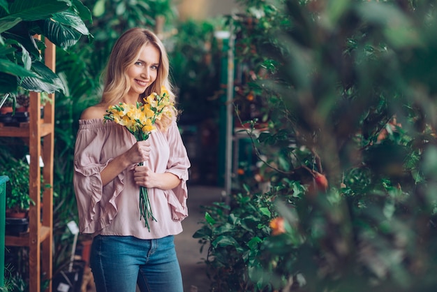노란 꽃 꽃다발을 들고 식물 종묘장에 서있는 젊은 여자를 웃 고