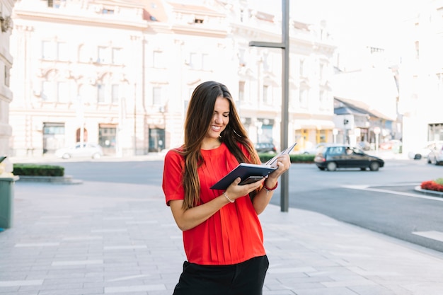 Smiling young woman standing on sidewalk reading diary