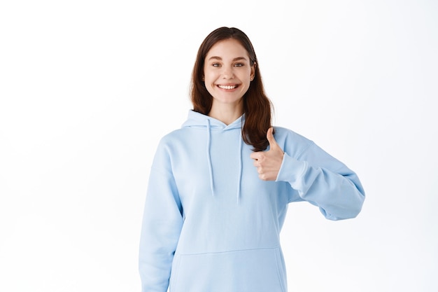 Smiling young woman stay positive, encourage you, give praises or guarantee, showing thumbs up in approval, standing against white wall