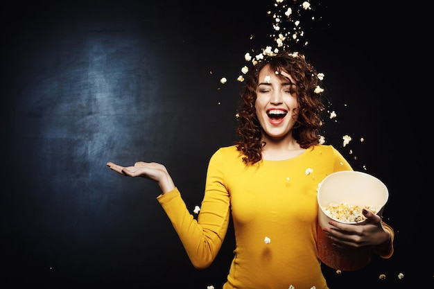 Smiling young woman staying under popcorn shower with right hand up