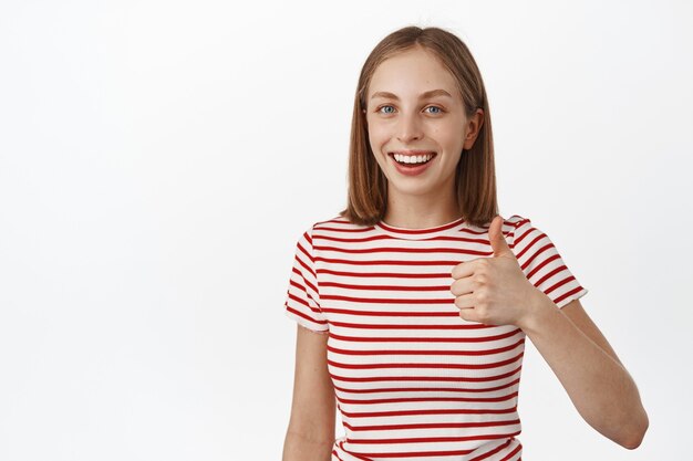Smiling young woman, student showing thumb up in approval, look satisfied, recommend product, like and agree, positive feedback, standing against white wall.