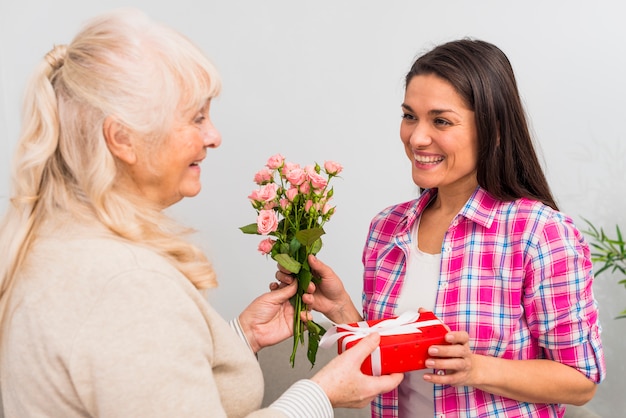 Free photo smiling young woman taking gift and rose bouquet from her senior woman