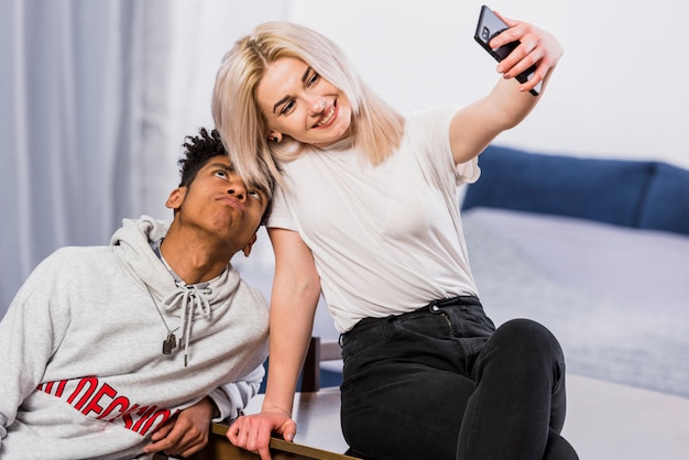 Smiling young woman taking selfie on mobile phone with his boyfriend making funny face