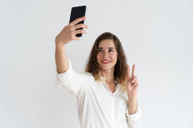 Улыбается молодая женщина, принимая selfie фото и показывая знак победы.