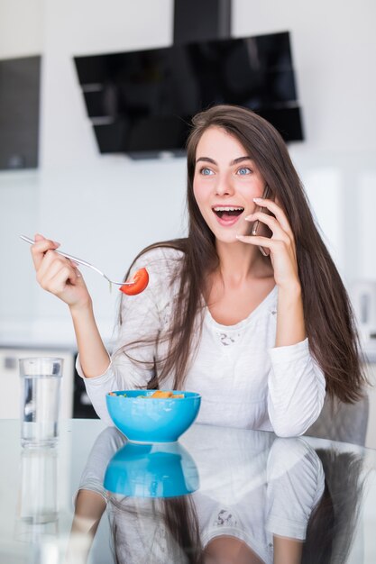 Smiling young woman talking on mobile phone while eat salad in a kitchen