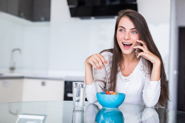 キッチンでサラダを食べながら携帯電話で話している若い女性を笑顔