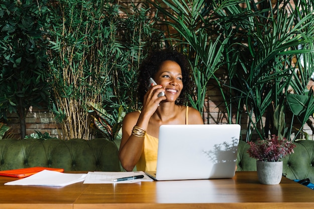 Smiling young woman talking on mobile phone with laptop; documents and pen on wooden table