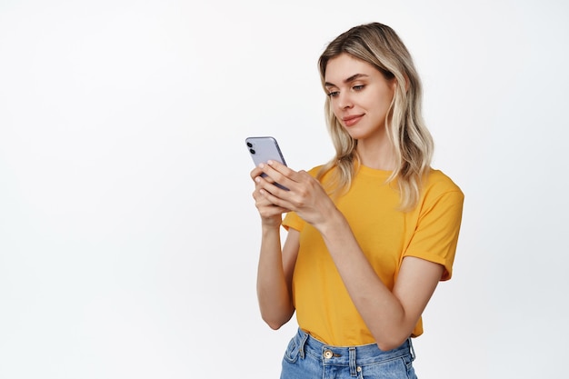 Smiling young woman using smartphone, looking at mobile phone with satisfied face expression, reading message, standing on white