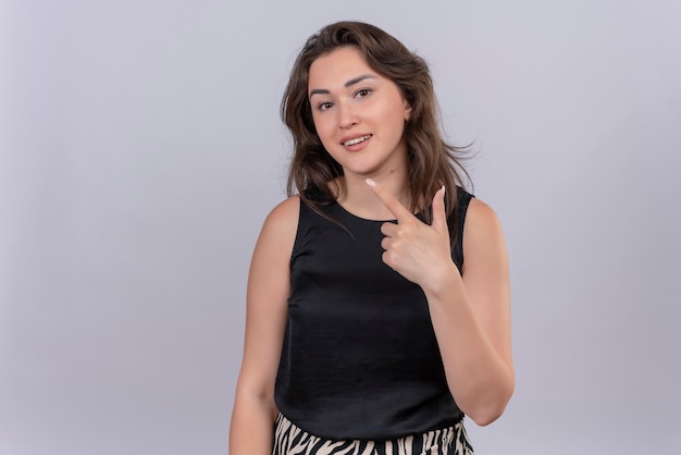 Smiling young woman wearing black undershirt points to up on white wall