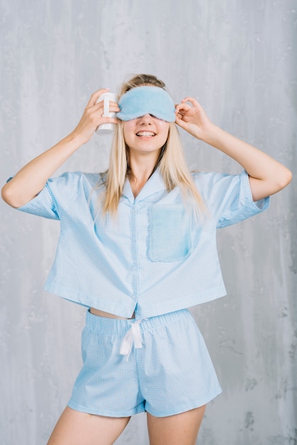 Giovane donna sorridente che indossa la maschera per gli occhi blu di sonno contro la parete