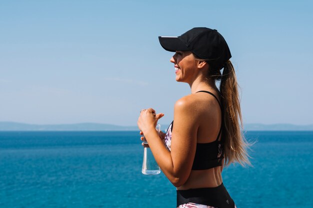 水の瓶を手にして、海の前に立っているキャップを着ている若い女性に笑顔