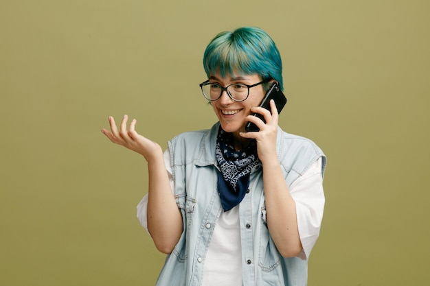 Smiling young woman wearing glasses bandana on neck looking at side talking on phone showing empty hand isolated on olive green background