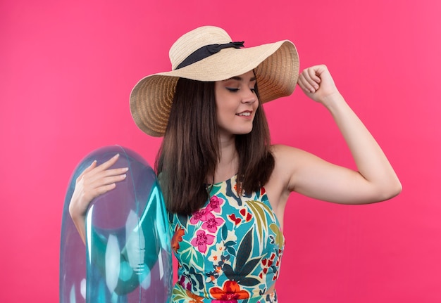 Smiling young woman wearing hat holding swim ring and lifting her hand on isolated pink wall