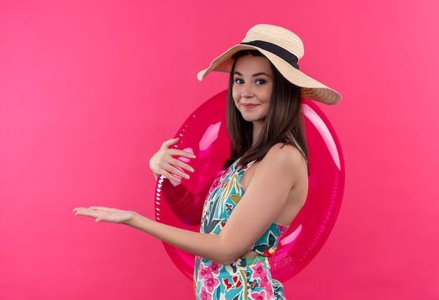 孤立したピンクの壁の左側にある水泳リングを押しながら手を指している帽子をかぶっている若い女性の笑みを浮かべてください。