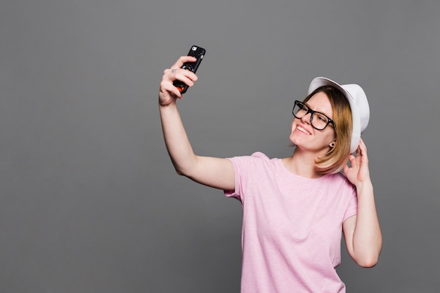 Smiling young woman wearing hat taking selfie on mobile phone against grey backdrop