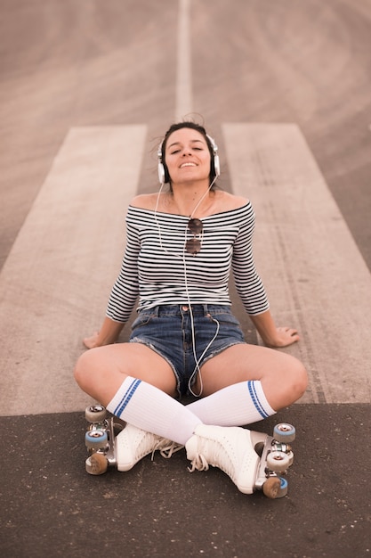 헤드폰에 도로 듣는 음악에 앉아 롤러 스케이트를 입고 웃는 젊은 여자