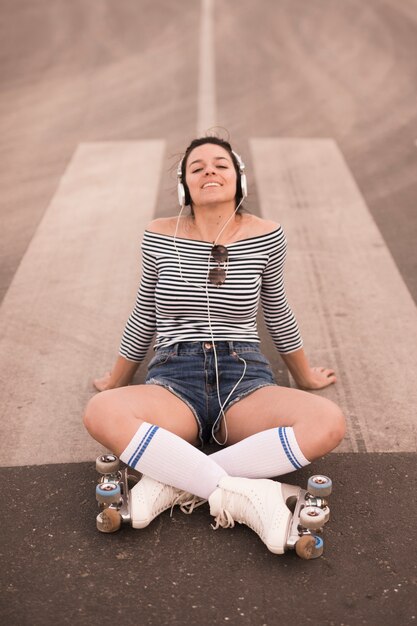 Усмехаясь коньки ролика молодой женщины нося сидя на музыке дороги слушая на наушниках