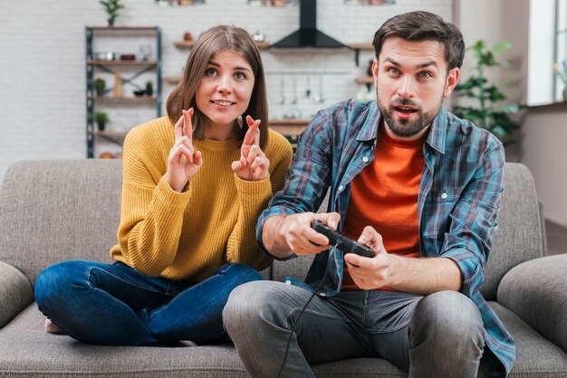 Smiling young woman with crossed fingers sitting near the man playing video game