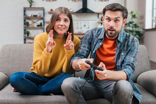 ビデオゲームをプレイする男のそばに座って交差指を持つ若い女性を笑顔