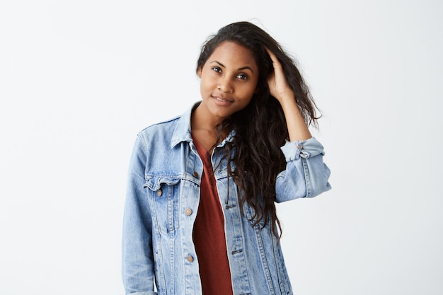 Smiling young woman with dark wavy hair in denim jacket and red t-shirt, posing and playing with her hair. Afro-American girl dressed casually cheerfully .