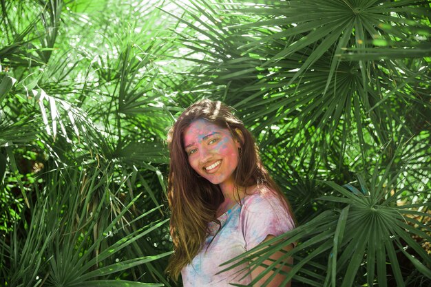 緑のヤシの葉の中で立っている彼女の体にホーリーカラーを持つ若い女性の笑みを浮かべてください。