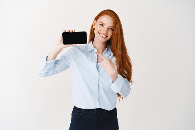 Улыбающаяся молодая женщина с рыжими волосами показывает пустой экран телефона, указывая на дисплей смартфона, стоящий на белом фоне