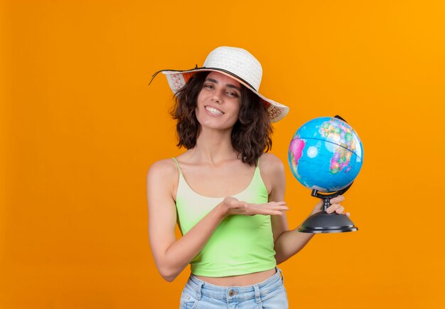A smiling young woman with short hair in green crop top wearing sun hat showing a globe 