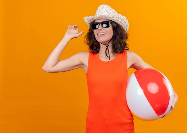 A smiling young woman with short hair in an orange shirt wearing sun hat and sunglasses holding inflatable ball showing goodbye with hand 