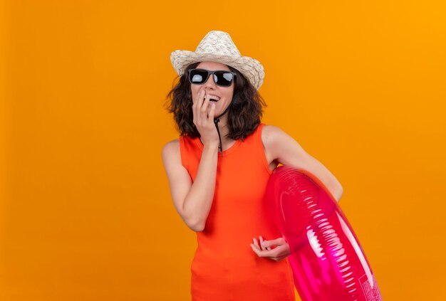 A smiling young woman with short hair in an orange shirt wearing sun hat and sunglasses holding inflatable ring keeping hand on face 