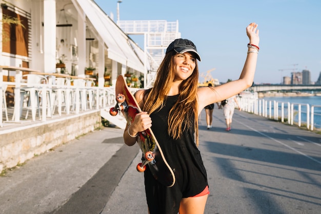 Улыбается молодая женщина с скейтборд, подняв руки во время прогулки по улице