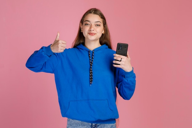 Smiling young woman with smartphone showing hand gesture with thumb up indoors