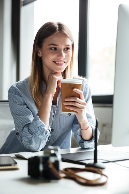 コーヒーを飲んでコンピューターを使用してオフィスで働く若い女性の笑みを浮かべてください。