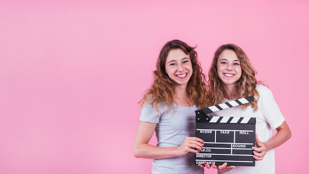 Free photo smiling young women holding clapper board in hands against pink backdrop