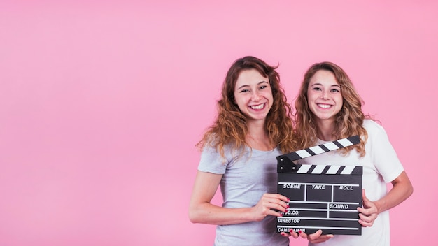 Smiling young women holding clapper board in hands against pink backdrop