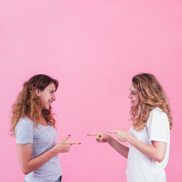 Free photo smiling young women pointing fingers to each other against pink backdrop