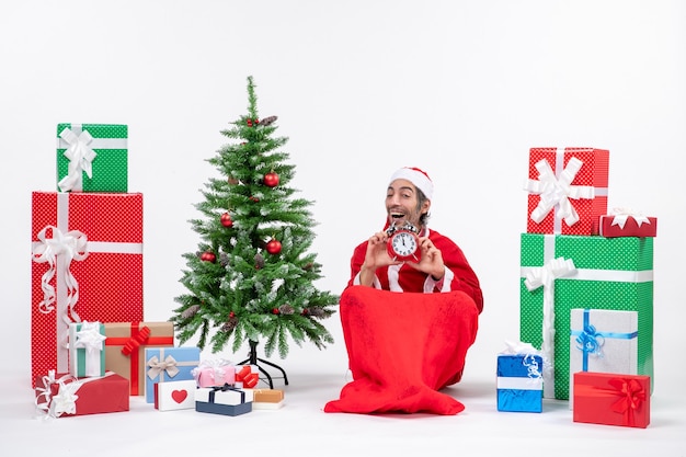 Smilinghappy satisfied young man celebrate new year or christmas holiday sitting on the ground and holding clock near gifts and decorated xsmas tree