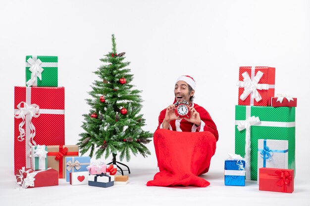 Smilinghappy satisfied young man celebrate new year or christmas holiday sitting on the ground and holding clock near gifts and decorated xsmas tree