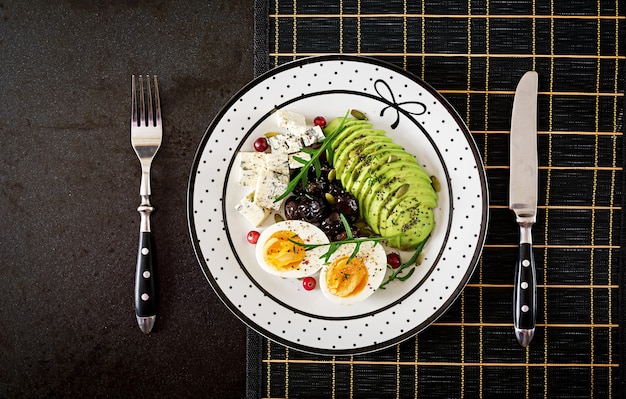スナックまたは健康的な朝食-黒い表面にブルーチーズ、アボカド、ゆで卵、オリーブのプレート。上面図