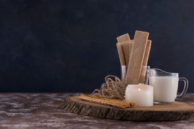 Закуски и крекеры со стаканом молока на деревянной доске с белой свечой в стороне.