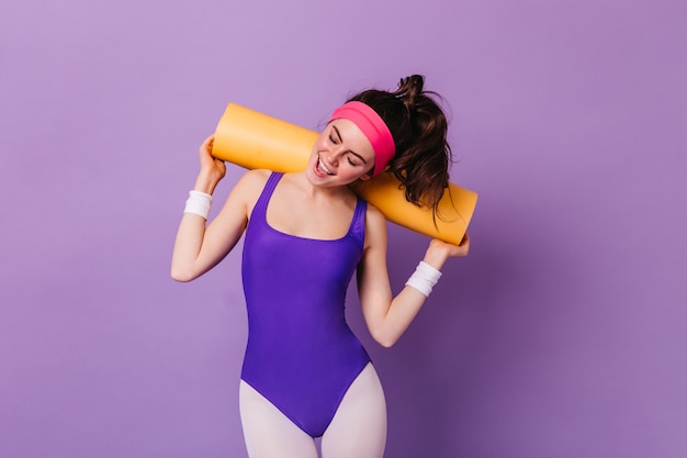 Снимок привлекательной женщины в спортивной одежде в стиле 80-х, позирующей с ковриком для аэробики на фиолетовой стене