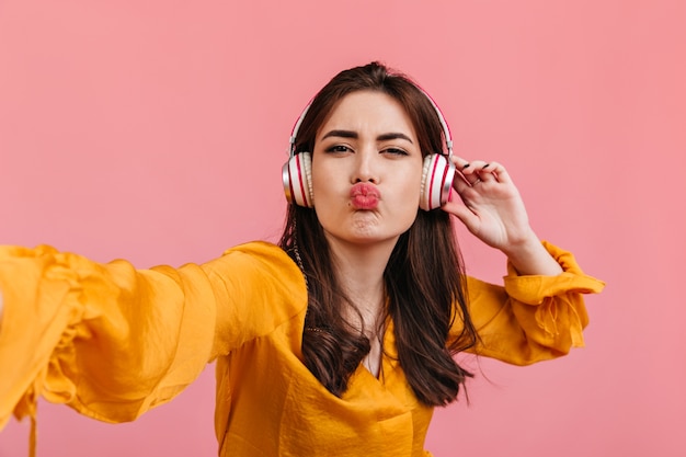 Snapshot of brunette in yellow outfit. Funny girl in headphones sends kiss, posing for selfie on pink wall.