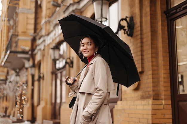 Snapshot of charming Asian girl in stylish trench coat holding black umbrella on city street