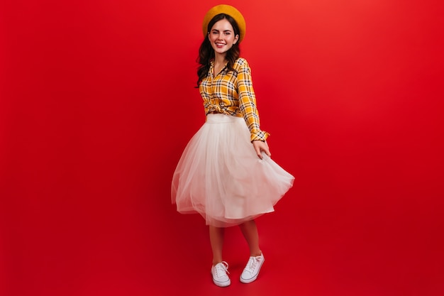 Snapshot full-length of curly dark-haired girl in bright outfit. Woman in beret and fluffy skirt is spinning on red wall.