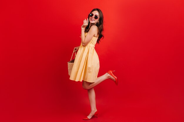 Snapshot full-length of pretty girl in short yellow dress on red wall. Woman with dark wavy hair in sunglasses is smiling cute.